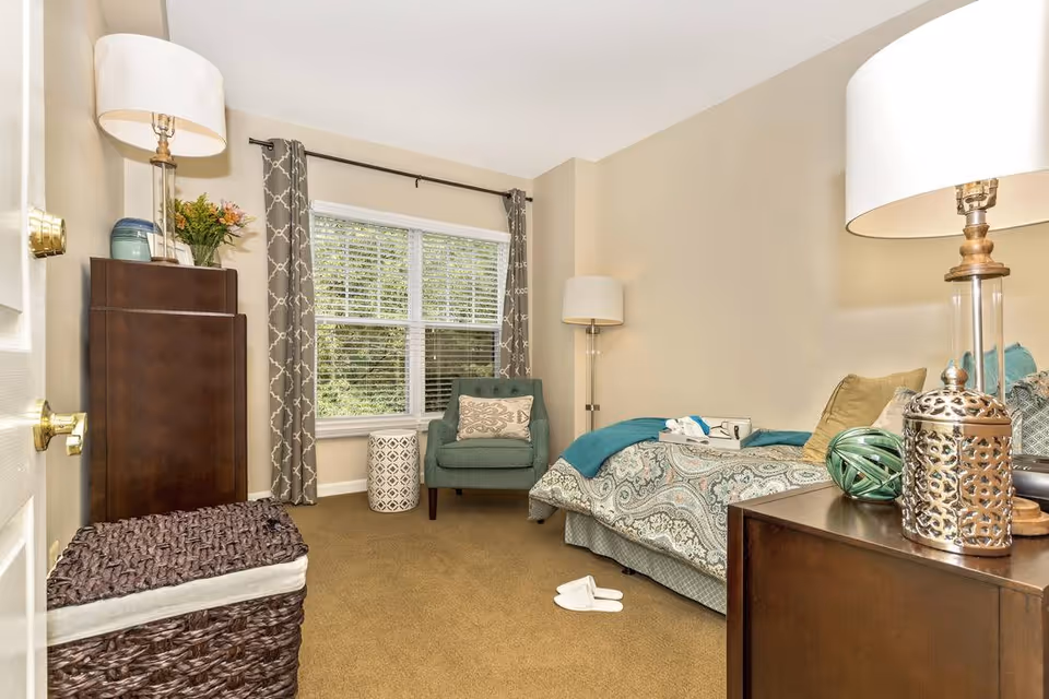 A cozy bedroom with a single bed covered in a patterned bedspread and multiple pillows. There is a teal armchair with a decorative pillow next to a window with patterned curtains. A small round side table is beside the chair. The room also features a dark wooden dresser with a lamp, decorative items, and a basket with a cushion on top. The walls are painted beige and the carpet is brown.