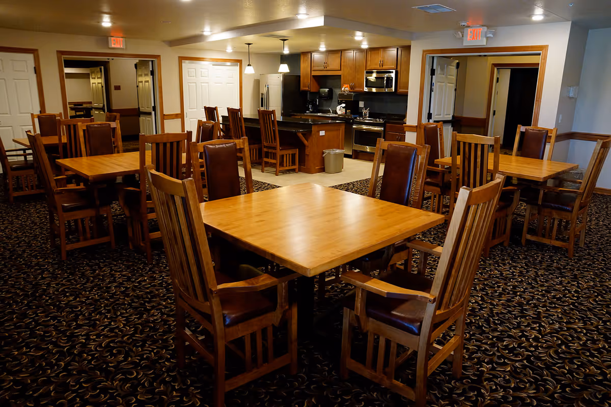 Carpeted dining room with wooden tables and chairs and a kitchen area in the background.