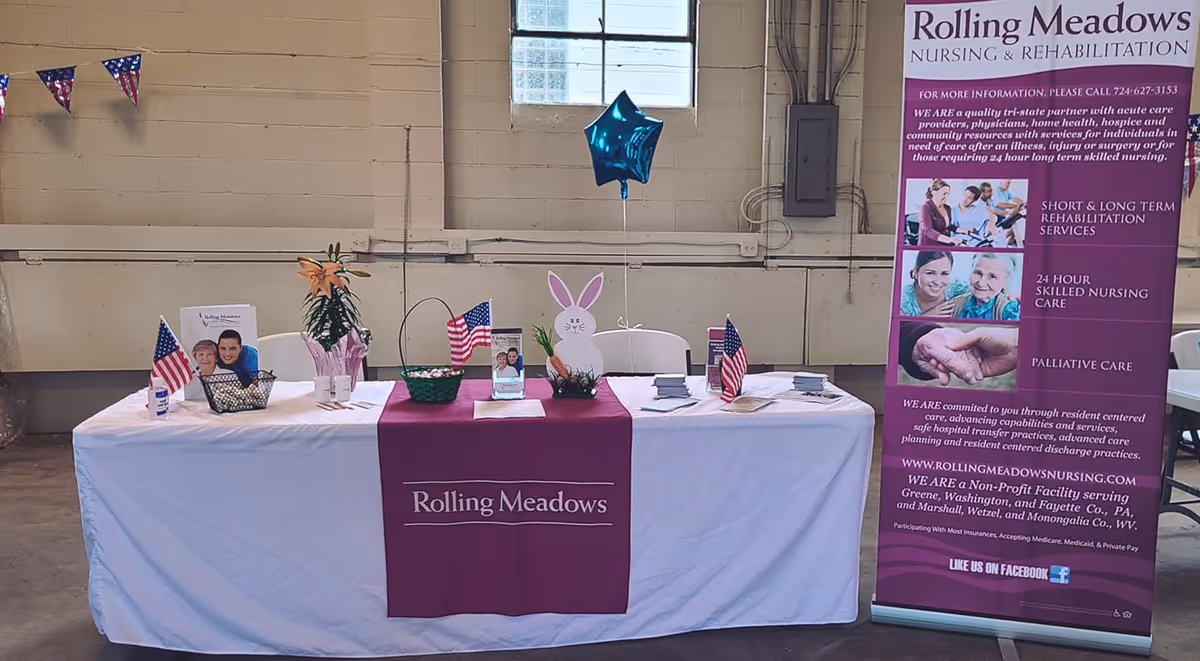 Indoor informational table for Rolling Meadows nursing & rehabilitation with brochures, American flags, a balloon, and a tall banner.