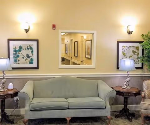 A cozy sitting area with a light green upholstered sofa centered between two wooden side tables, each holding a decorative lamp. Above the sofa are two framed botanical prints and a rectangular mirror reflecting a hallway with additional framed artwork. The walls are painted a soft beige with white trim, and the lighting is warm and inviting.