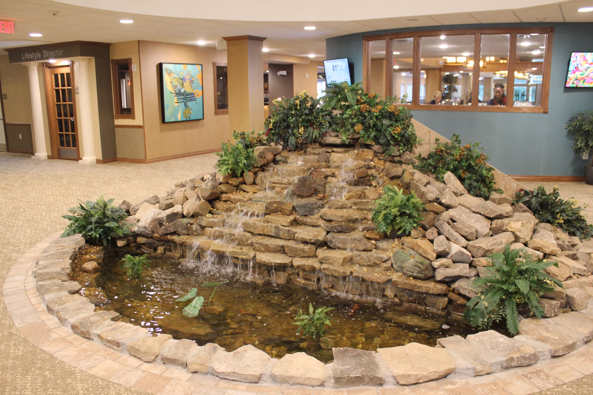 Indoor water feature with cascading waterfall made of rocks surrounded by plants in a carpeted common area of a retirement community. There is a sign for the Lifestyle Director office and a painting on the wall in the background.