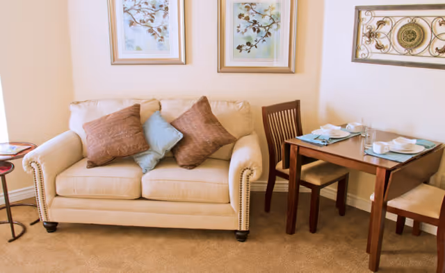 A cozy living area featuring a beige loveseat with three decorative pillows in brown and blue. Next to the loveseat is a small round side table. Adjacent to the loveseat is a wooden dining table set for two with white dishes, cups, and blue placemats. The walls are decorated with two framed floral paintings and a decorative metal wall art piece.