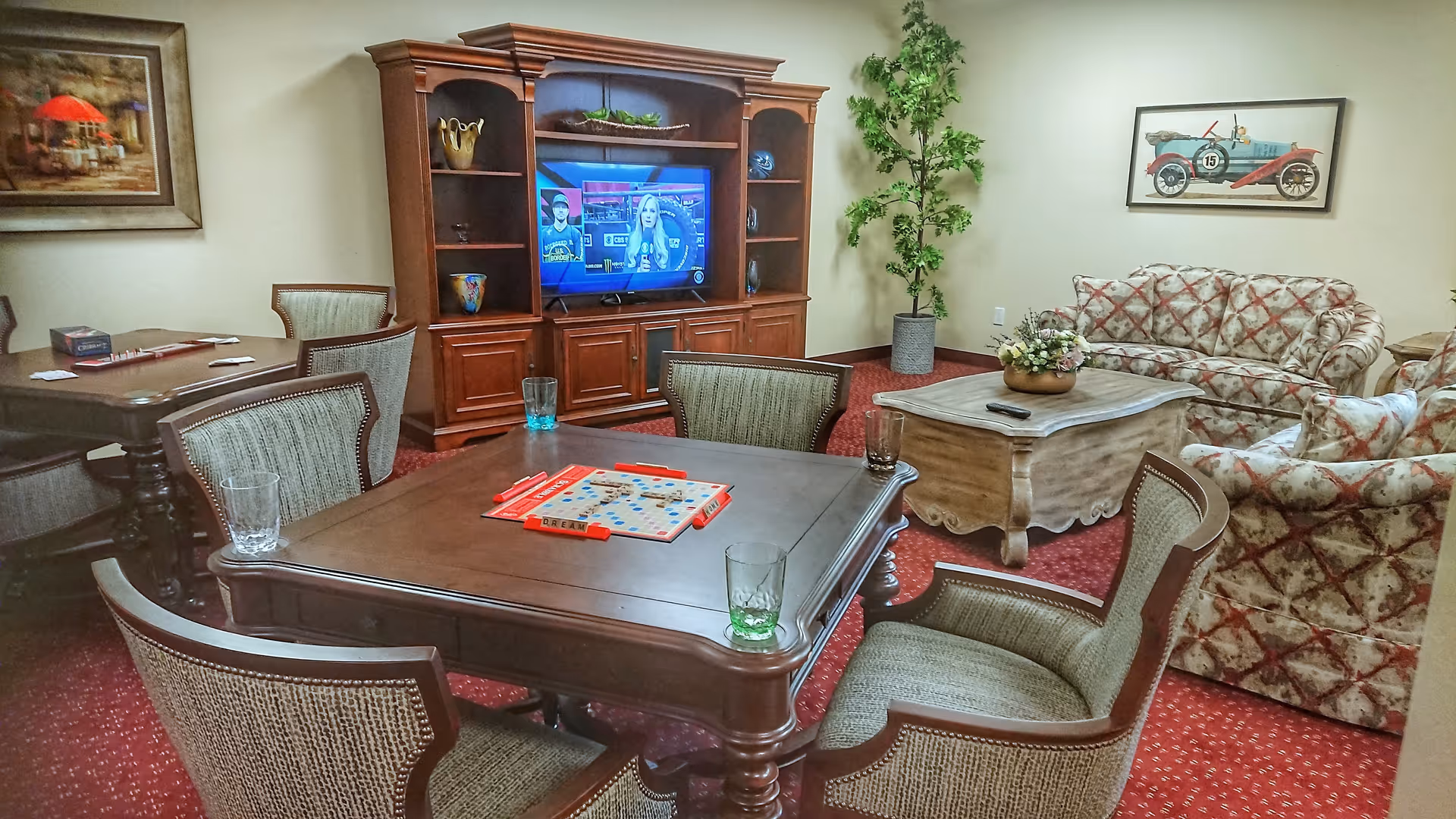 A cozy common room with a wooden entertainment center holding a TV, a coffee table with a floral arrangement, patterned sofas, and a table with chairs set up for a board game. The room has red carpet and framed artwork on the walls.