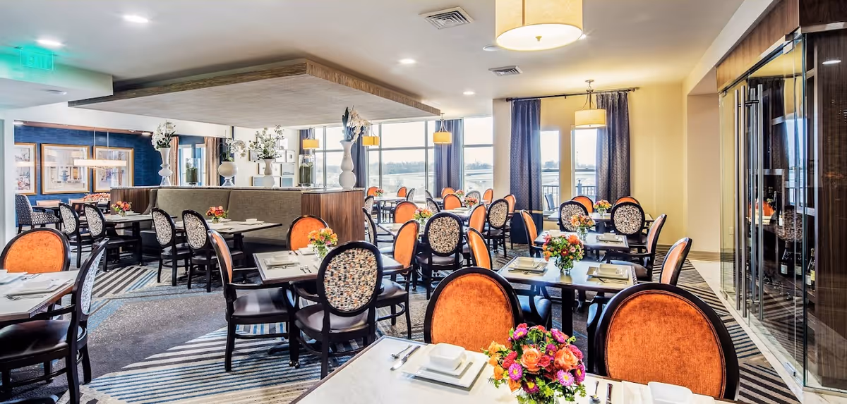 A bright and elegant dining room with multiple tables set with white plates, silverware, and colorful floral centerpieces. The chairs have orange and patterned upholstery. Large windows with curtains allow natural light to fill the space, and decorative vases with flowers are placed on a central divider. The room has modern lighting fixtures and a patterned carpet.