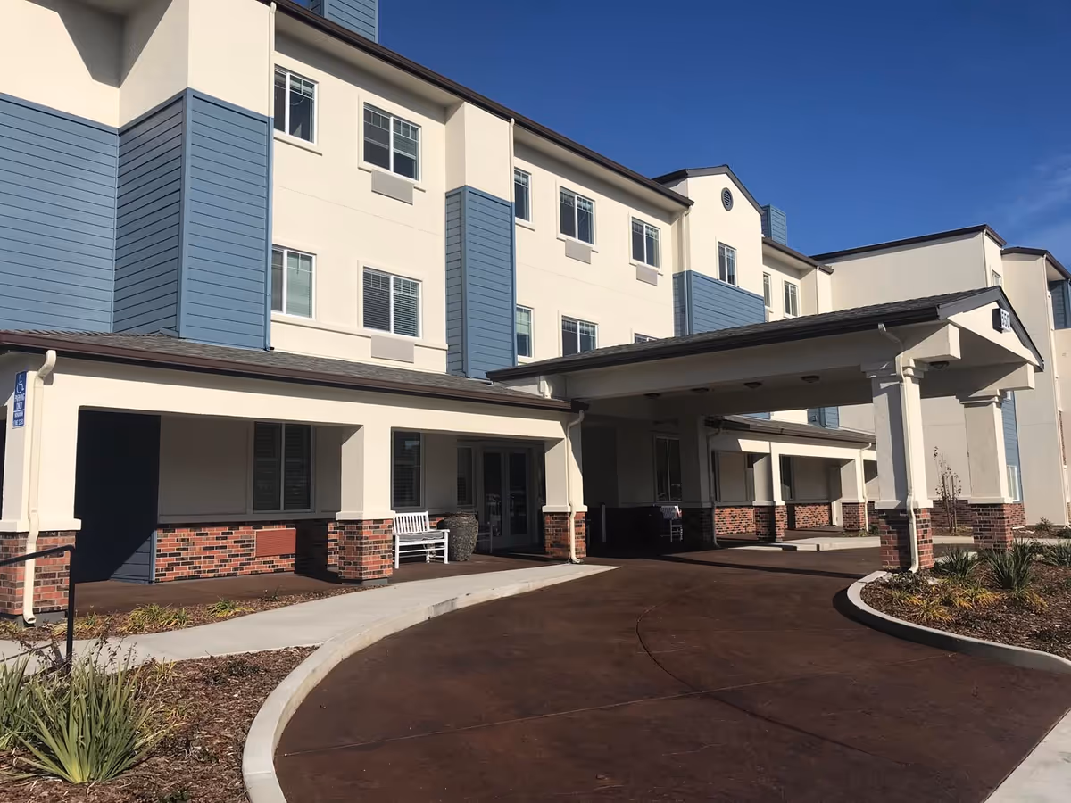 Exterior view of a three-story senior living facility building with a covered entrance driveway, white and blue walls, brick accents, and a clear blue sky.