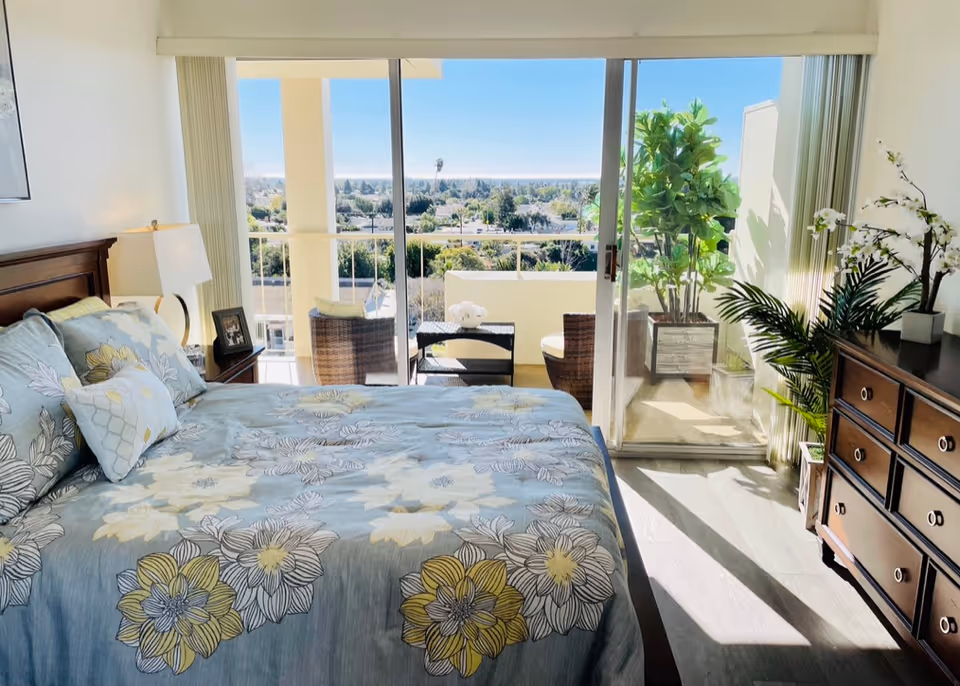 A bright bedroom with a large bed covered in a floral patterned bedspread in shades of blue, yellow, and white. To the left of the bed is a nightstand with a lamp and a framed photo. The room has a sliding glass door leading to a balcony with outdoor seating and plants. The balcony overlooks a scenic view of trees and a clear blue sky. On the right side of the room is a wooden dresser with plants and decorative flowers on top.