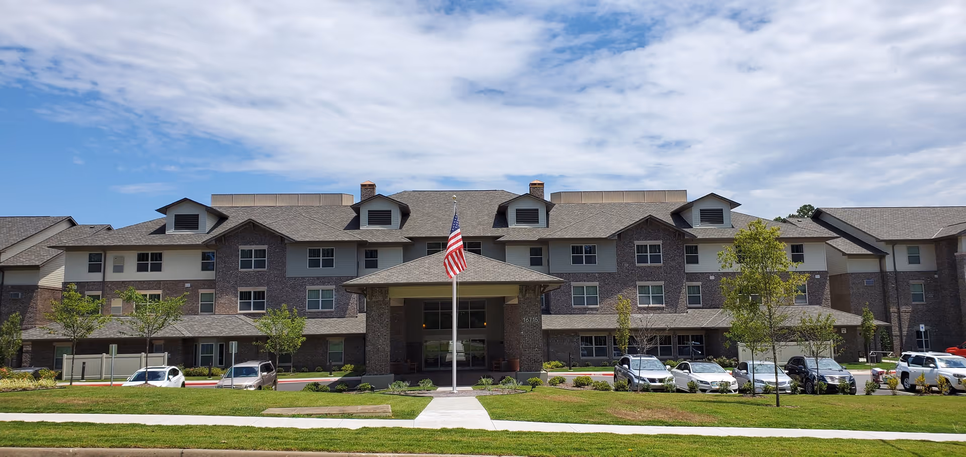 Front exterior view of Chenal Pines Retirement Resort, a large three-story building with a covered entrance, multiple windows, and an American flag on a flagpole in front. Several cars are parked along the driveway, and there are small trees and green lawns surrounding the building under a partly cloudy sky.