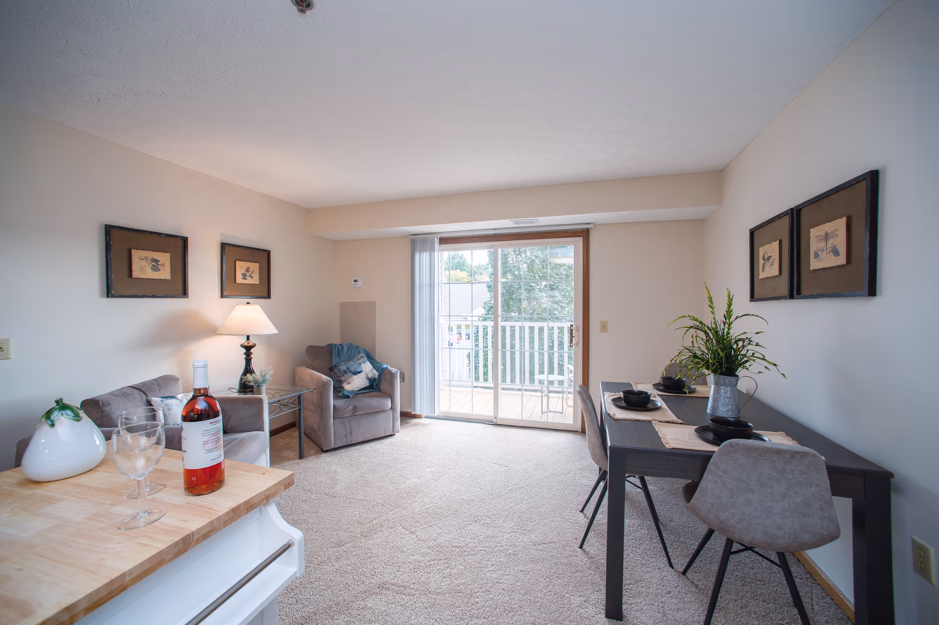 A bright living and dining area with beige carpet and light-colored walls. The room features a sliding glass door leading to a balcony with outdoor seating. There is a small dining table set with four chairs and place settings, a couple of armchairs with cushions, a side table with a lamp, and a kitchen counter with a bottle of wine and two wine glasses. Framed artwork hangs on the walls.
