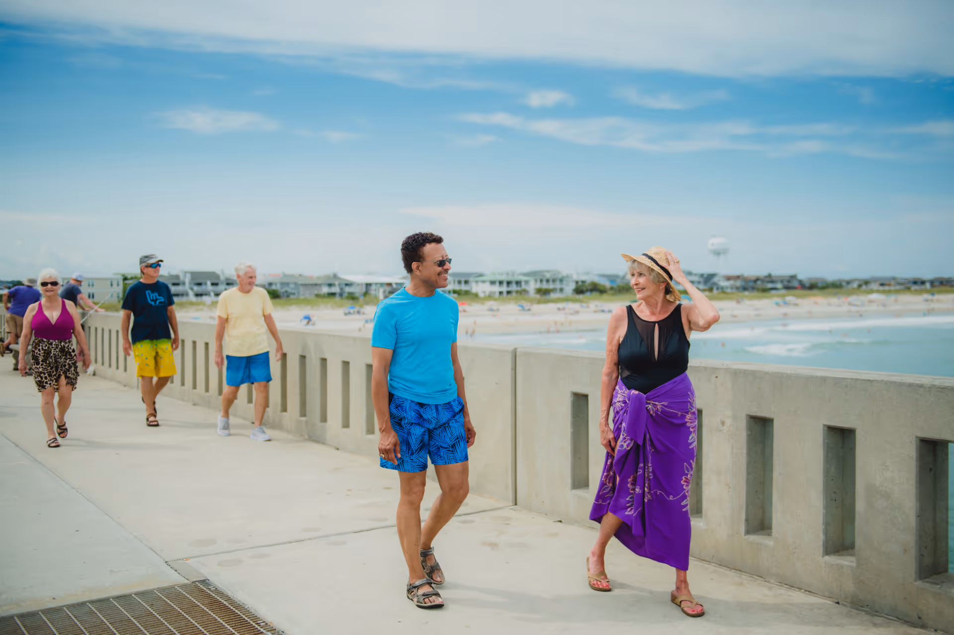 A group of older adults walking along a concrete boardwalk by the beach on a sunny day. Two people in the foreground are engaged in conversation, with the woman wearing a black top and purple sarong and the man wearing a blue shirt and blue patterned shorts. Other people walk behind them, and the beach and ocean are visible in the background under a partly cloudy sky.