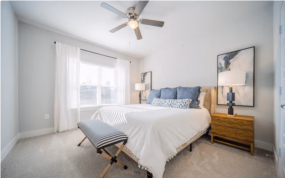 Bright, minimalist bedroom with a large bed topped with blue pillows, bedside tables and lamps, a striped bench at the foot, and a ceiling fan.