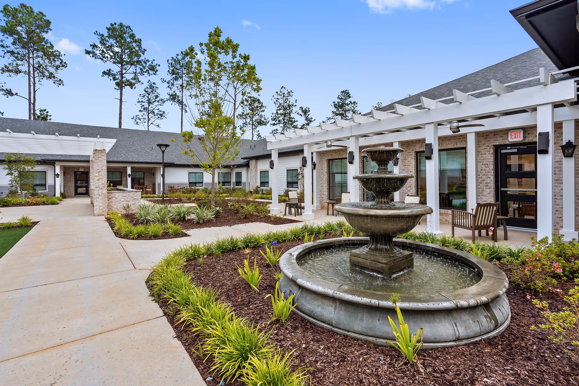 Outdoor courtyard featuring a multi-tiered fountain, landscaped paths, and seating under a pergola in front of a single-story building.