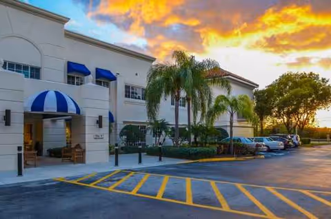 Facility exterior of Banyan Place with palm trees, parking lot, and blue-striped awning.