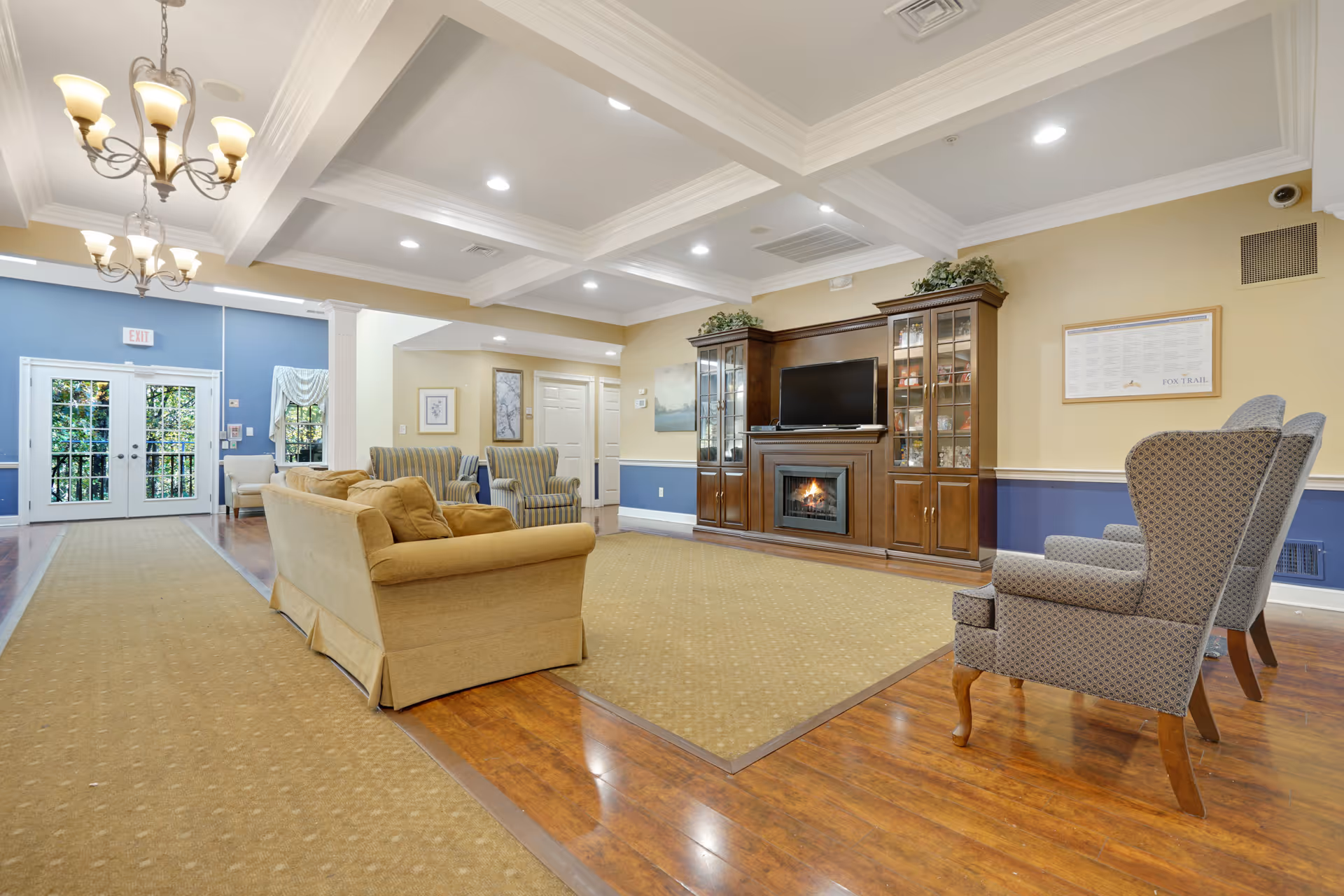 A spacious living room area in a senior living facility featuring a beige sofa, two patterned armchairs, and two striped armchairs arranged around a large area rug. The room has a wooden floor, coffered ceiling with recessed lighting, and two chandeliers. A wooden entertainment center with a fireplace and a TV is against the wall, with decorative plants on top. Double glass doors lead outside, and the walls are painted in soft yellow and blue tones.