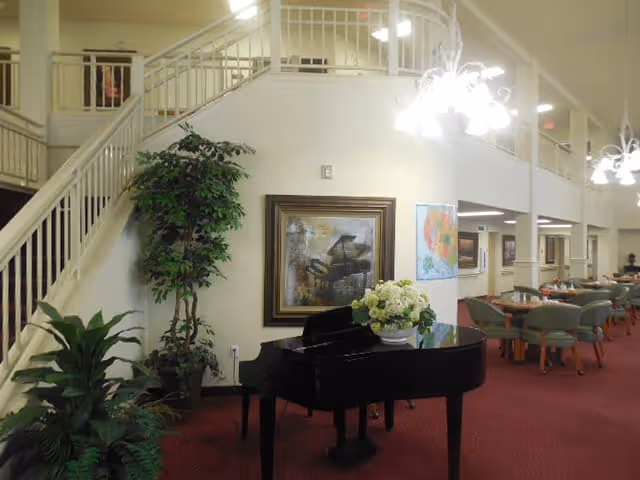 Interior view of a senior living facility common area featuring a black grand piano with a floral arrangement on top, potted plants, a staircase with white railings, framed artwork on the wall, and a dining area with tables and chairs in the background.