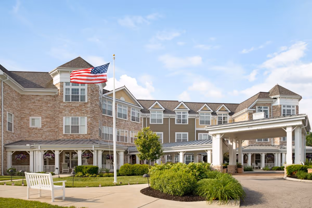 Exterior view of a senior living facility building with a stone and beige siding facade, multiple windows, and a covered entrance. An American flag is flying on a flagpole in front of the building, surrounded by well-maintained landscaping including bushes, a tree, and a white bench on a paved walkway under a partly cloudy sky.