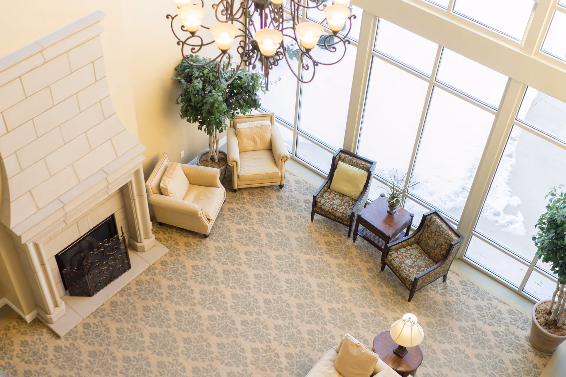 A bright living room area with large floor-to-ceiling windows letting in natural light. The room features a beige patterned carpet, a white stone fireplace with a decorative screen, two beige armchairs, two floral-patterned chairs with a small wooden table between them, a beige sofa with a side table and lamp, and two large potted plants. A chandelier hangs from the ceiling above.
