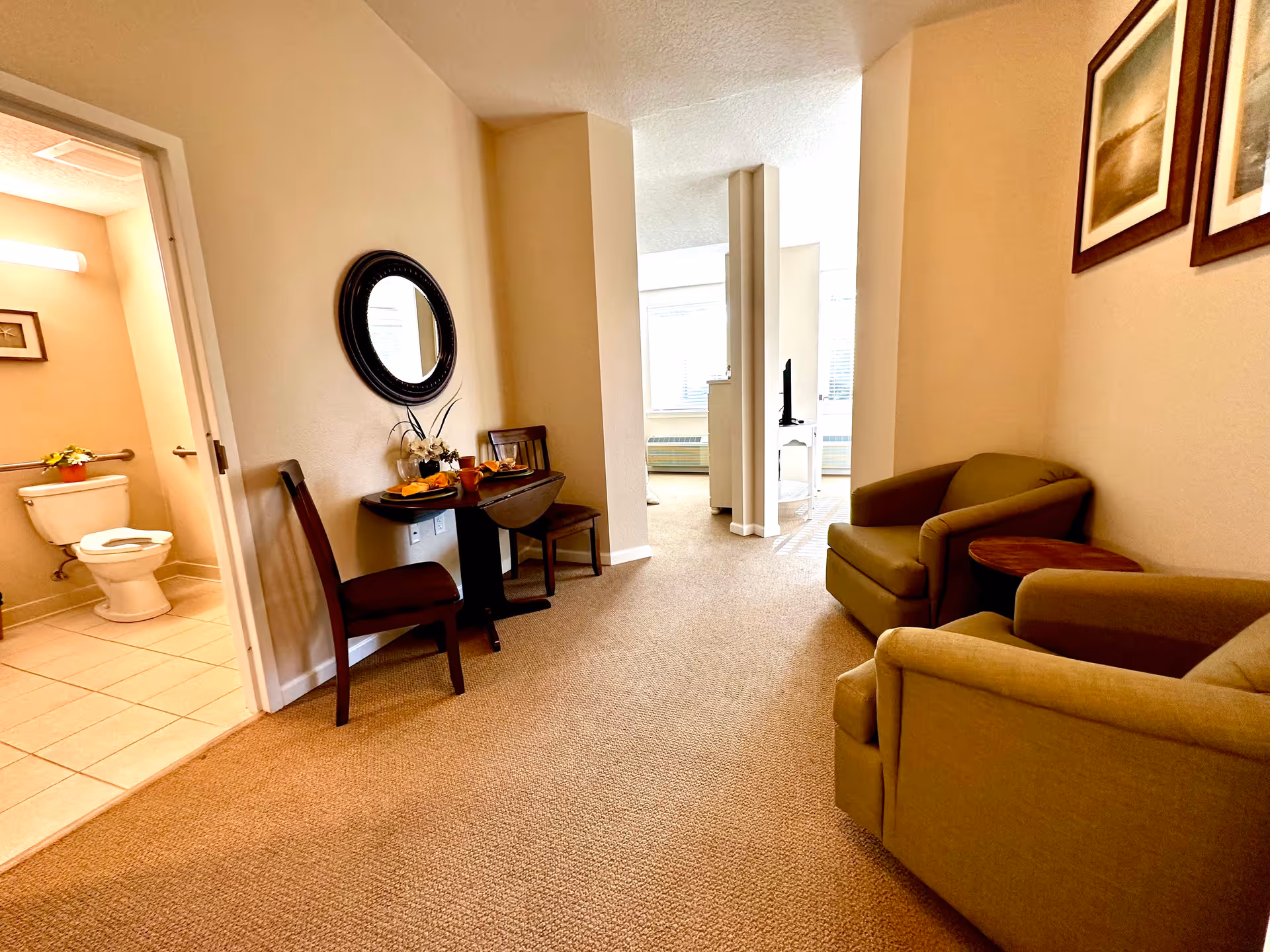 Small senior living suite interior with two armchairs, a small table and chairs, and a bathroom visible through an open door.
