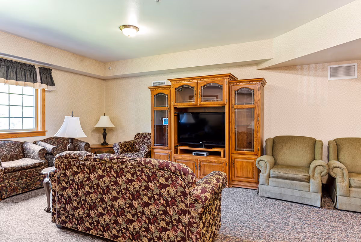 A cozy living room with floral patterned sofas and two green armchairs arranged around a wooden entertainment center holding a flat-screen TV. There are two table lamps on side tables near the sofas, a window with a valance on the left wall, and beige wallpaper with a subtle pattern.