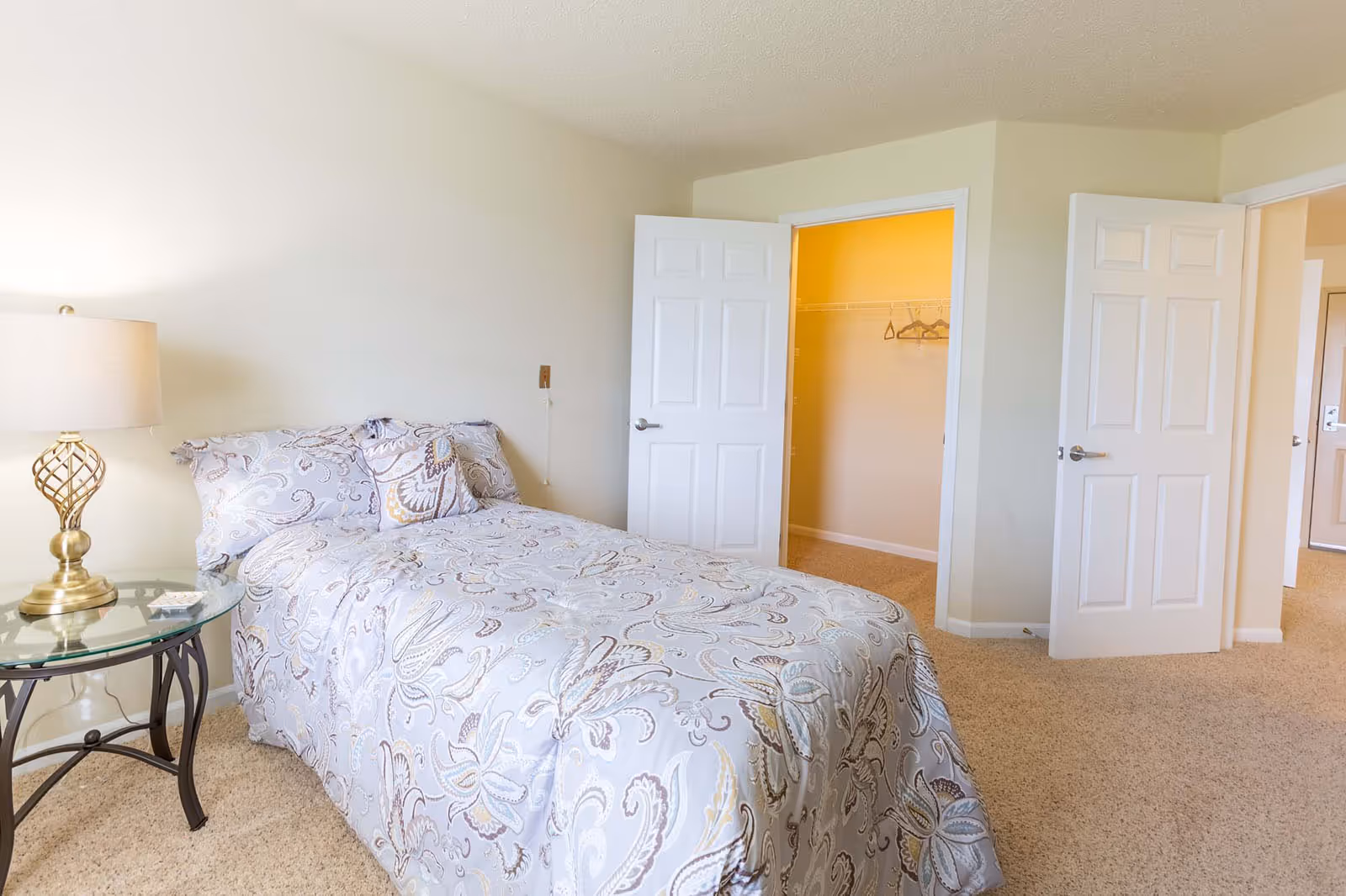 Sunlit bedroom with a single bed dressed in patterned bedding, a glass side table with a lamp, and open closet and room doors.