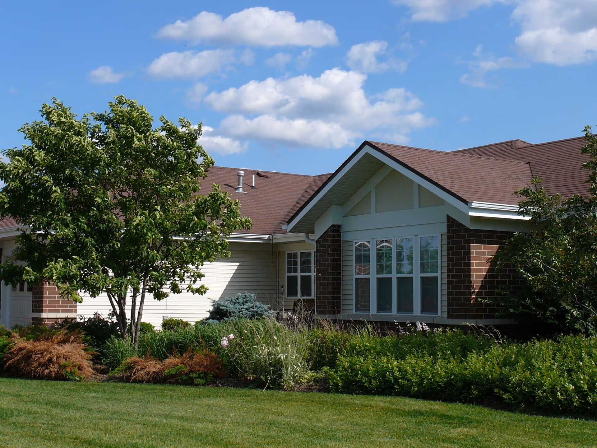 Exterior view of a single-story building with a brown roof and brick and siding walls, surrounded by green shrubs, trees, and a well-maintained lawn under a blue sky with scattered clouds.