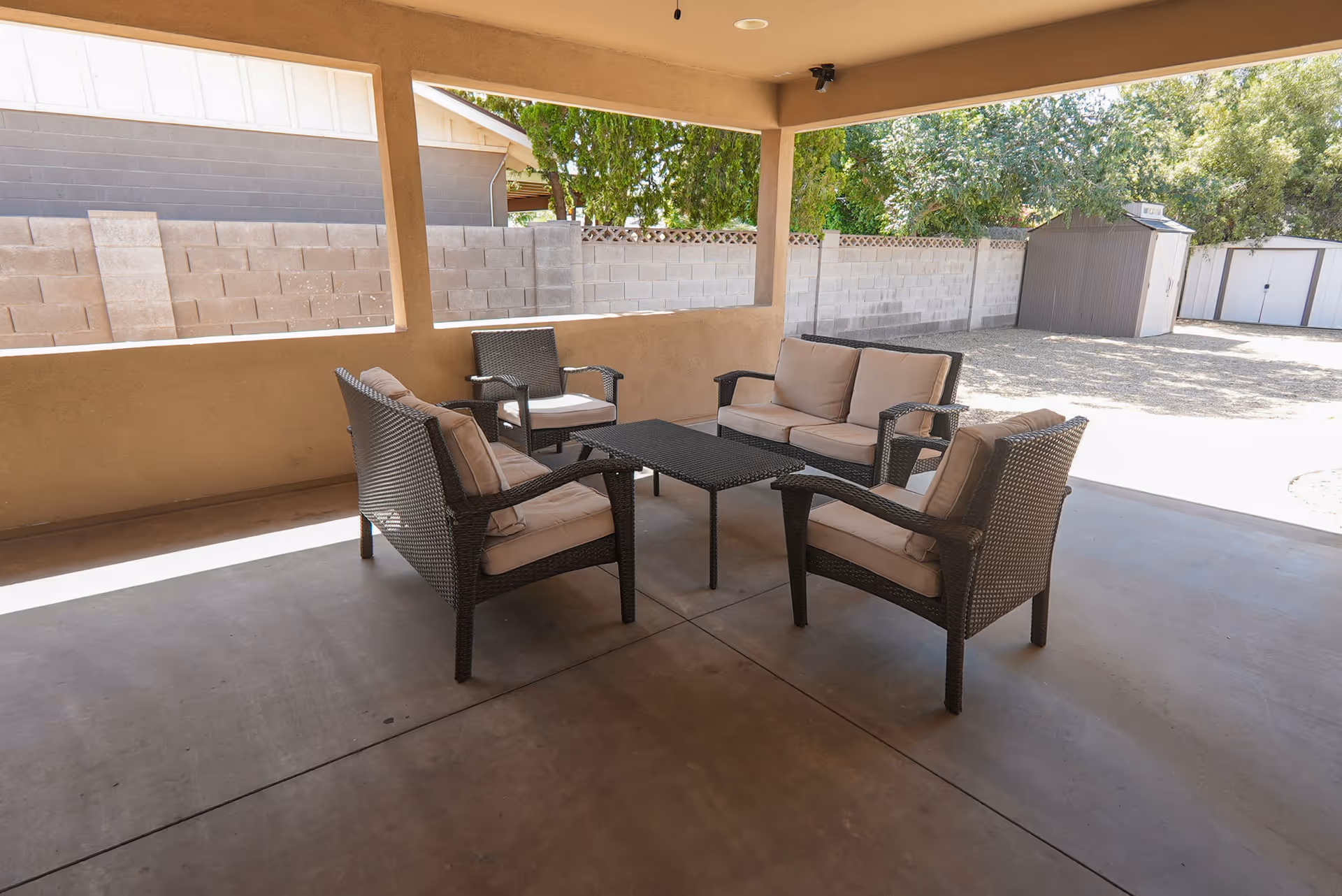 Covered outdoor patio with wicker seating and cushions arranged around a central coffee table on a concrete floor, with a cinderblock wall and storage sheds in the background.