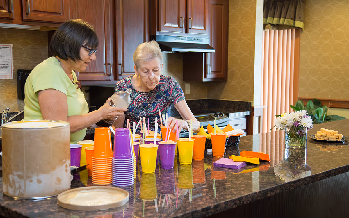 Two women in a senior living kitchen preparing colorful drinks on a large granite countertop with plastic cups and napkins.