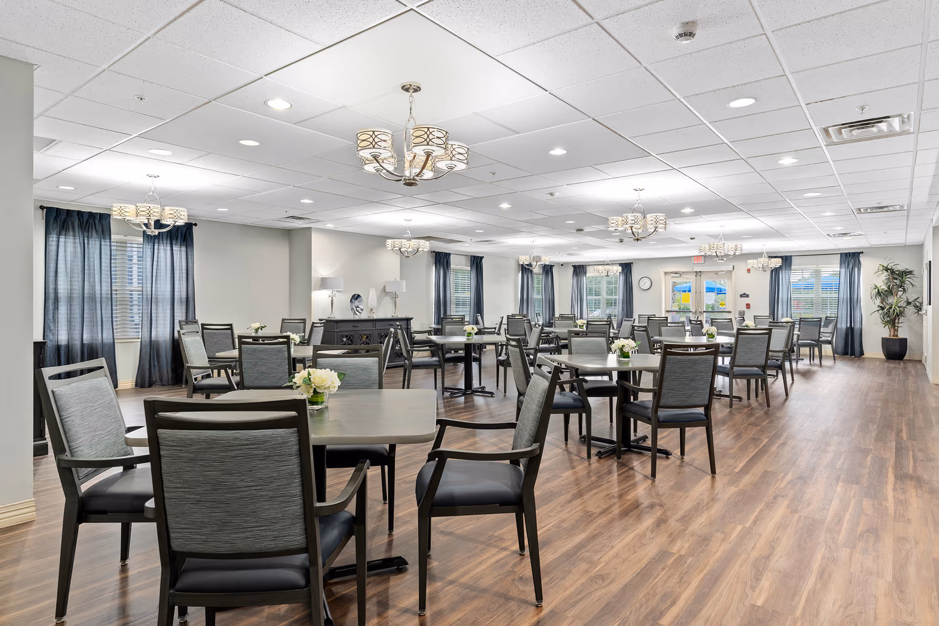 A spacious dining room with multiple tables and chairs arranged neatly. The room has wooden flooring, white walls, and several windows with dark curtains allowing natural light to enter. Modern chandeliers hang from the ceiling, and small flower arrangements are placed on each table. There is a sideboard with lamps and decorative items along one wall.