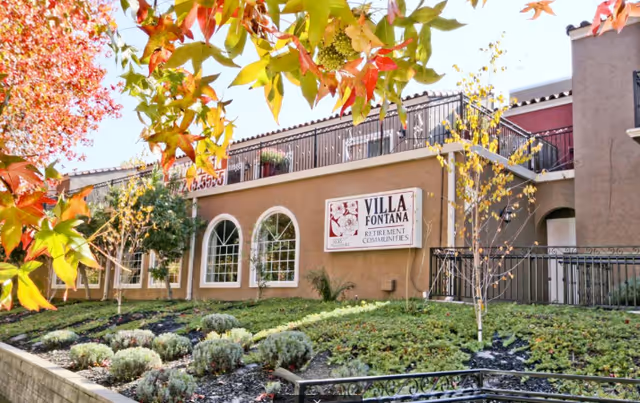 Exterior view of Villa Fontana Retirement Community building with beige walls, arched windows, and a sign displaying the community name. The foreground features a landscaped garden with various shrubs and trees with autumn-colored leaves.