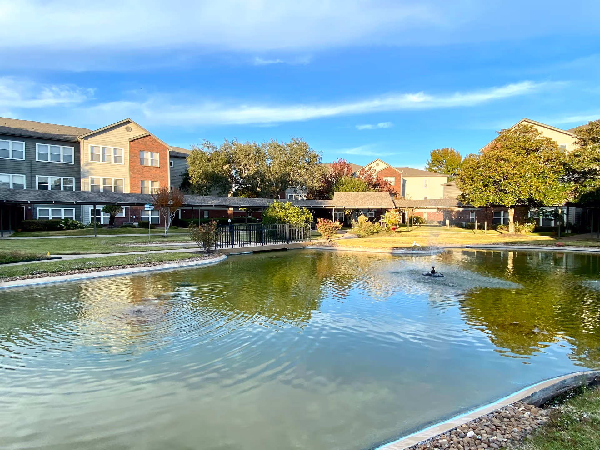 A serene outdoor area of a senior living facility featuring a large pond with a small fountain in the center, surrounded by well-maintained grass, trees, and shrubs. In the background, there are multi-story residential buildings with a mix of brick and siding exteriors under a blue sky with some clouds.