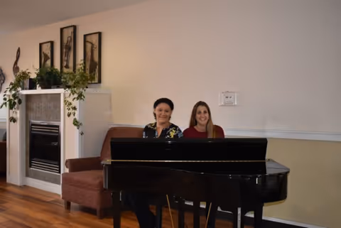 Two women sitting behind a black grand piano in a room with wooden flooring. Behind them is a brown armchair, a white fireplace with green plants on the mantel, and three framed pictures on the wall above the fireplace.