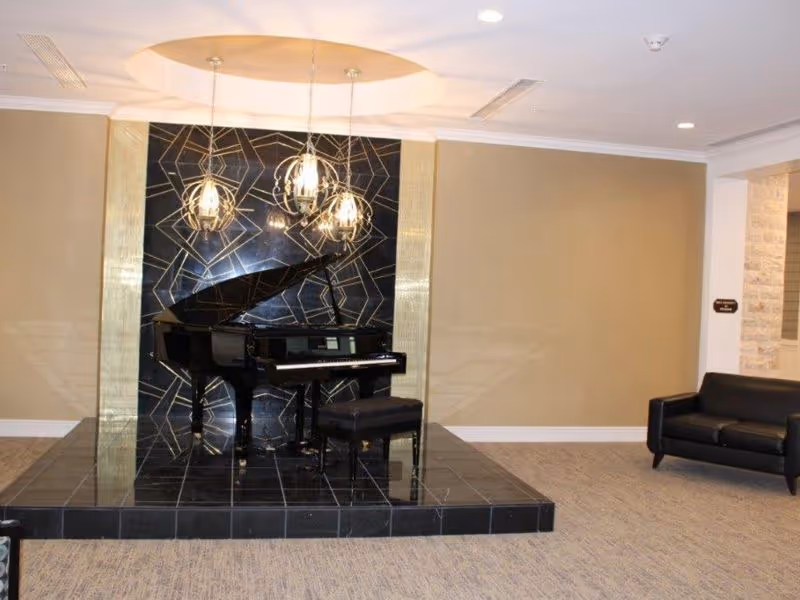 A black grand piano with a matching bench is displayed on a raised black tiled platform in a room. Behind the piano is a decorative black and gold geometric patterned wall panel. Three modern pendant lights hang above the piano. To the right, there is a black leather loveseat against a beige wall. The floor is carpeted in a neutral tone.