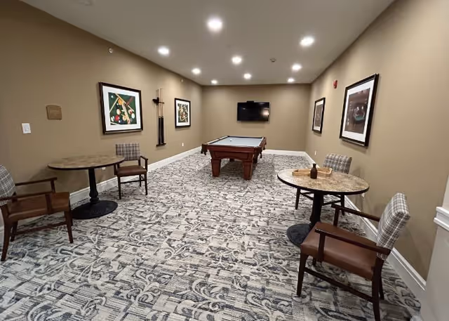Carpeted recreation room with a pool table, wall-mounted TV, framed art, and small round tables with chairs.