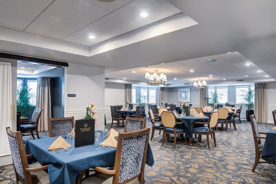 A spacious dining room with multiple tables covered in blue tablecloths, each set with beige napkins, glassware, and floral centerpieces. The room features patterned carpet, large windows with curtains, and elegant chandeliers hanging from the ceiling.