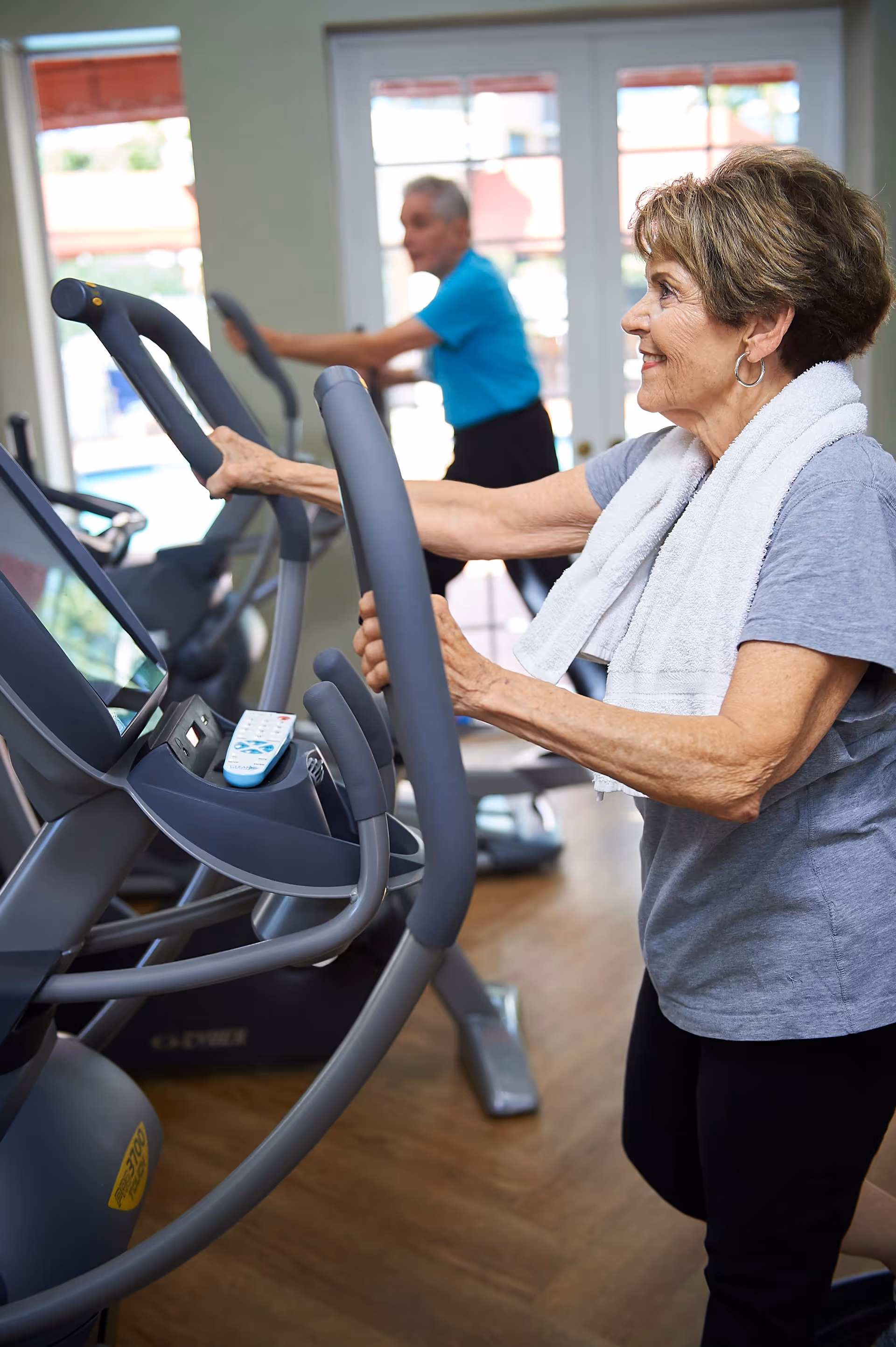 Two elderly individuals exercising on elliptical machines in a fitness room with wooden floors and large windows letting in natural light. One woman in the foreground has a white towel around her neck and is smiling.