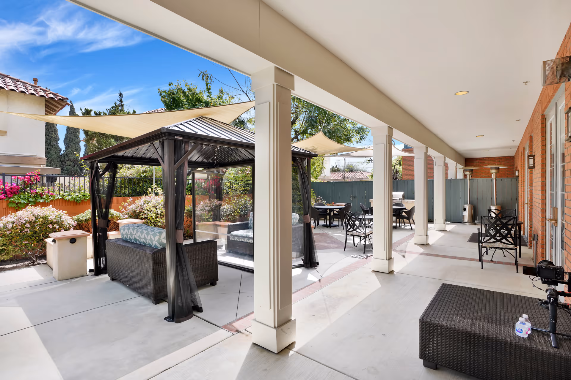 Outdoor patio area at Kirkwood Orange featuring a covered seating area with cushioned chairs under a gazebo, several tables and chairs for dining or socializing, shade sails overhead, and surrounding greenery and flowers.