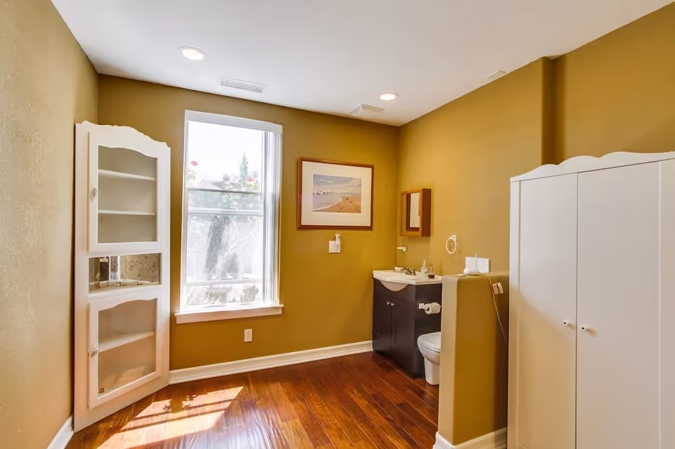 A bathroom with mustard yellow walls and wooden flooring. There is a white cabinet with glass doors on the left side near a window letting in natural light. On the right side, there is a white wardrobe and a small sink with a mirror above it. A toilet is partially visible behind a half wall next to the sink. A framed picture hangs on the wall above the sink.