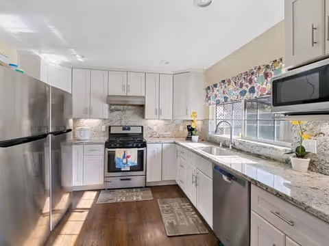 Bright kitchen with white cabinets, stainless steel refrigerator, stove, and dishwasher. Granite countertops and backsplash with a window above the sink featuring a floral valance. Wooden floor with two mats, one near the stove and one near the sink.