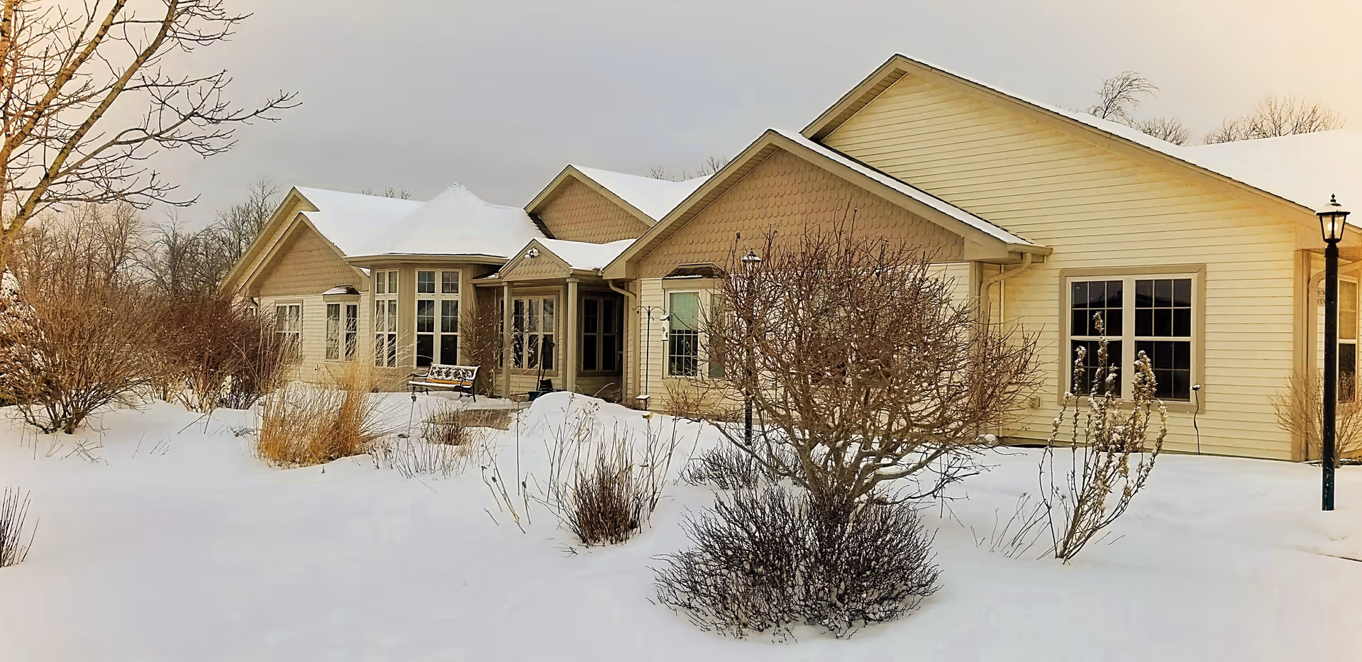 Exterior view of a single-story senior living facility named Forest View Manor, surrounded by snow-covered ground and leafless bushes and trees, with a bench near the entrance and a lamppost on the right side.