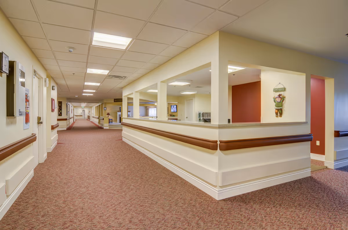 A wide, carpeted hallway in a healthcare facility with beige walls and ceiling tiles. The hallway features handrails along the walls and a reception or nurse station area with open windows on the right side. The station area has a countertop and a decorative scarecrow hanging on the wall. The hallway is well-lit with ceiling lights and extends into the distance with doors and framed pictures along the walls.