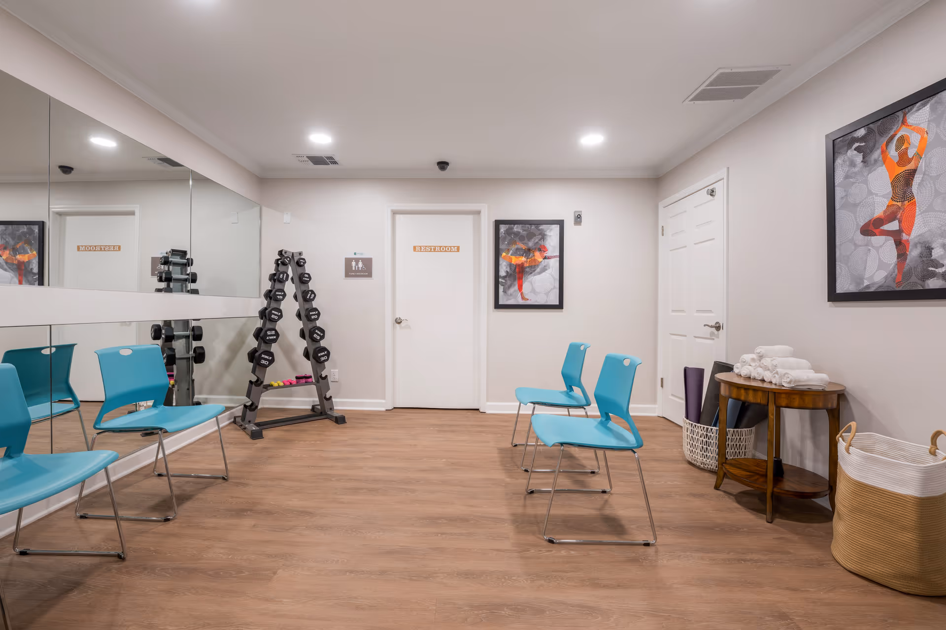 A small exercise room with four turquoise chairs arranged in two rows facing a wall with a large mirror. There is a rack of dumbbells in the corner next to a door labeled 'Restroom'. On the right side, there is a small wooden table with rolled white towels and baskets containing yoga mats and other items. The walls are light-colored and decorated with framed artwork of a person in a yoga pose.