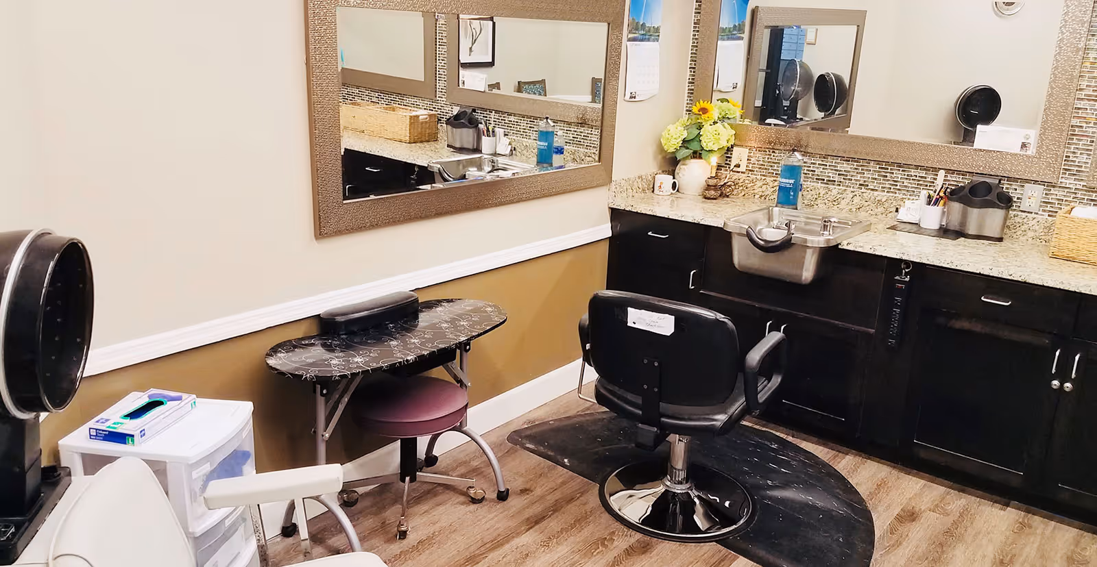 Interior view of a hair salon or beauty station in a senior living facility, featuring a black salon chair on a protective mat, a small table with a stool, a hair dryer hood, a countertop with a sink, a large mirror, and various hair care items including a bottle of sanitizer and a flower vase.