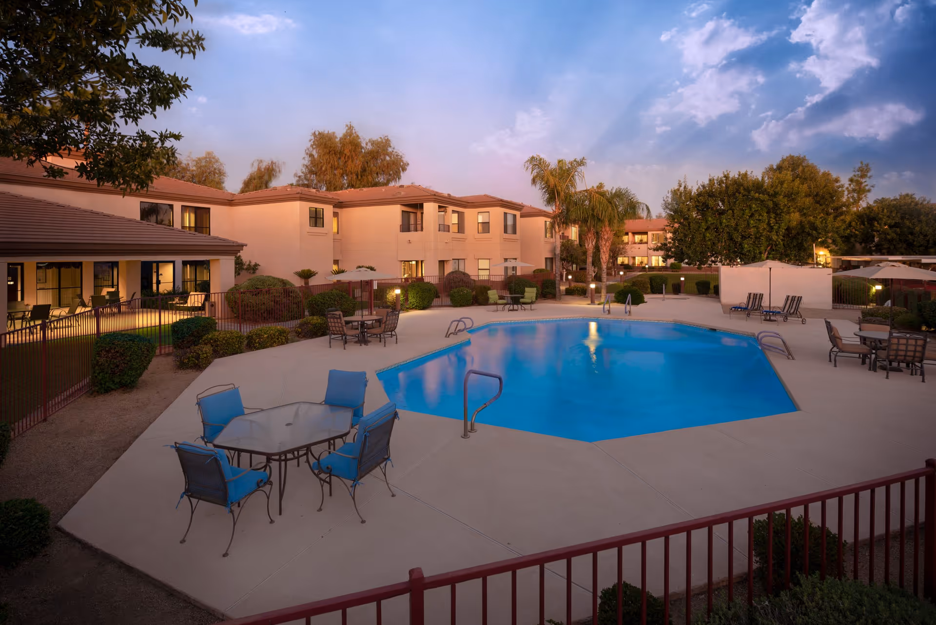 Outdoor swimming pool area at Desert Winds Assisted Living with patio tables and chairs with blue cushions, surrounded by a red fence. The pool is octagonal and there are palm trees, bushes, and a two-story building in the background under a partly cloudy sky at dusk.