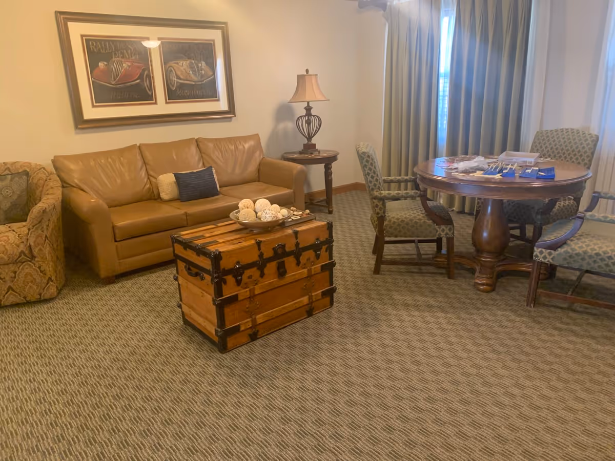 Cozy sitting area with a leather sofa, wooden trunk coffee table, round dining table and upholstered chairs near a curtained window.