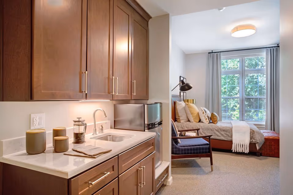 A cozy studio apartment featuring a small kitchenette with wooden cabinets, a sink, a mini refrigerator, and countertop accessories. Adjacent to the kitchenette is a bedroom area with a bed adorned with multiple pillows, a side table with a lamp, a comfortable chair, and a large window with curtains letting in natural light.