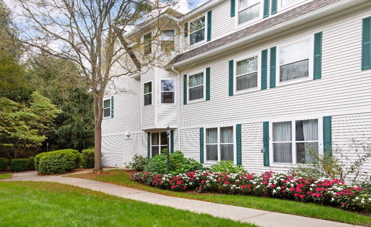 Exterior view of a white multi-story building with green shutters and multiple windows. There is a concrete walkway in front of the building, bordered by a well-maintained lawn and flower beds with red and white flowers. Trees and shrubs are visible around the building.