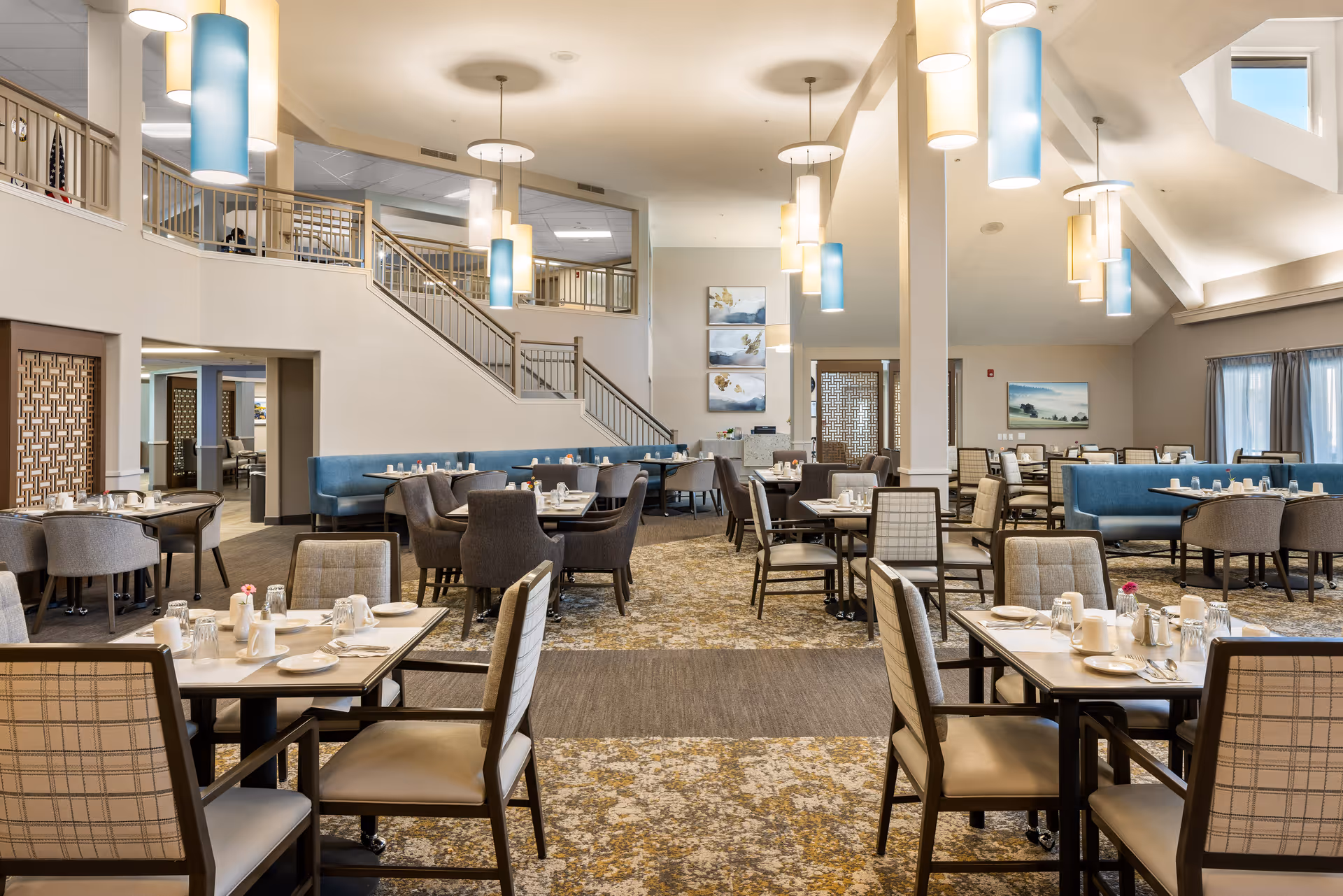 A spacious and well-lit dining room in a senior living community featuring multiple tables set with plates, cups, and utensils. The room has a high ceiling with modern pendant lights in blue and white, comfortable upholstered chairs, and blue cushioned bench seating along the walls. There is a staircase leading to an upper level and decorative artwork on the walls.