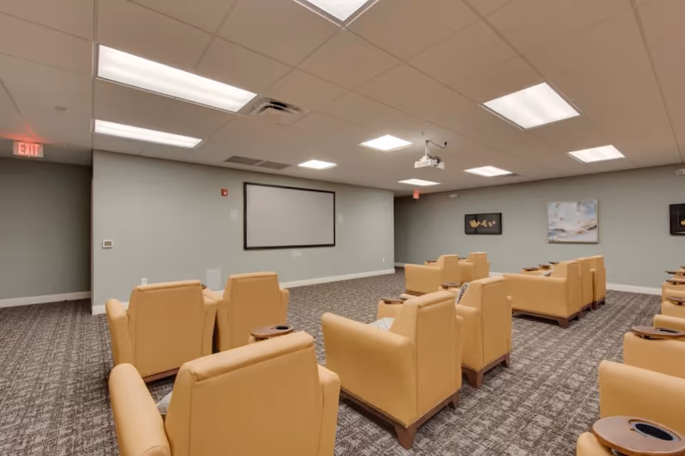 A media room with rows of tan reclining chairs facing a projector and blank screen.