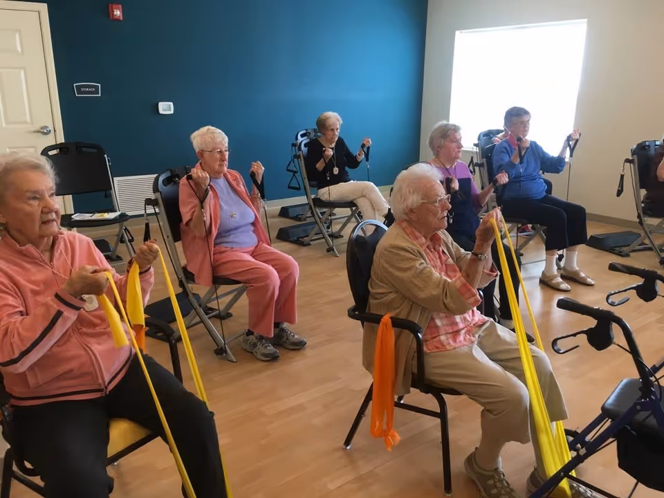 A group of elderly women seated in chairs in a room with wooden floors and a teal accent wall, participating in a seated exercise class using yellow resistance bands.