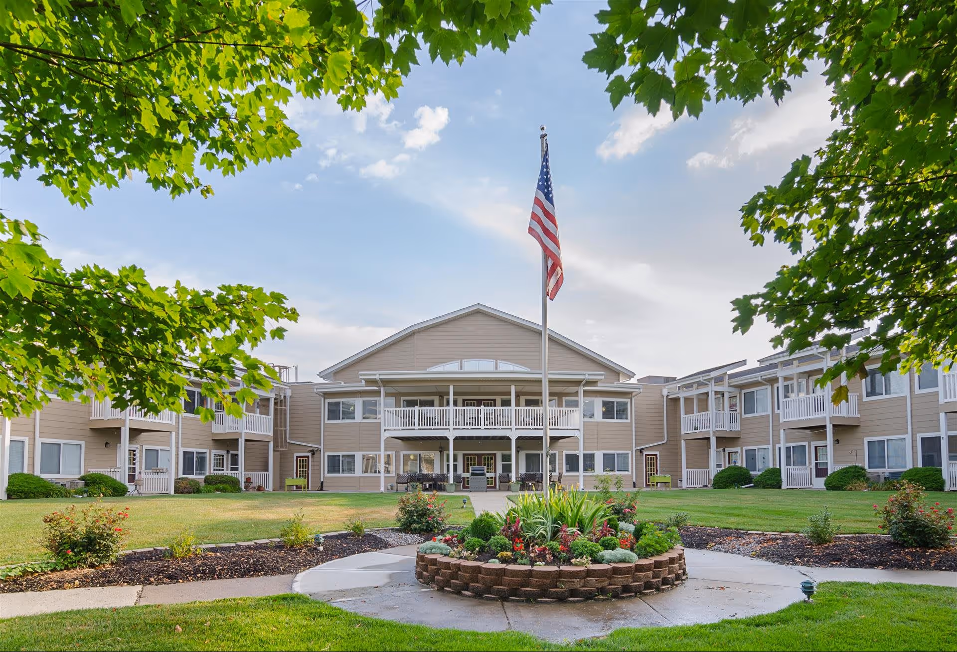 Exterior view of The Gardens at Barry Road Assisted Living and Memory Care facility showing a two-story building with balconies, a central flagpole with an American flag, and a landscaped garden area with green grass and trees framing the scene.
