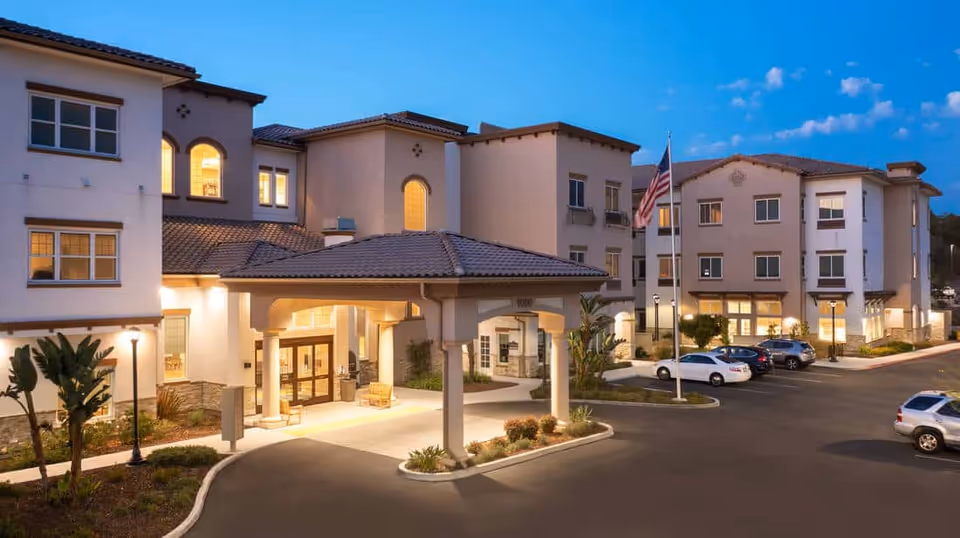 Exterior front entrance of a multi-story senior living building with a covered porte-cochère, illuminated windows, landscaping, and parked cars at dusk.